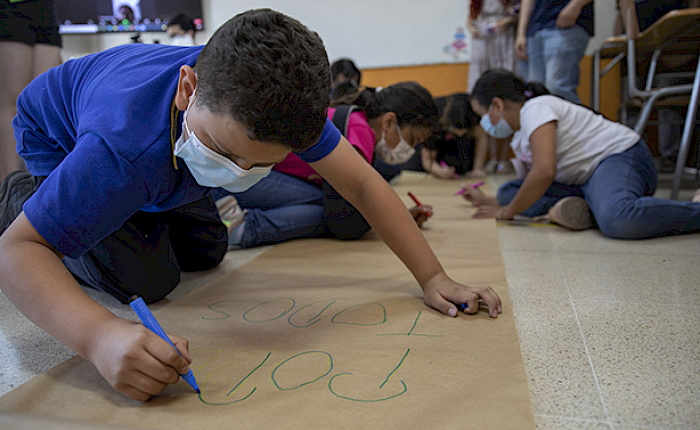 Primer plano de niño escribiendo sobre cartulina, en el suelo. En segundo plazo hay otros niños.