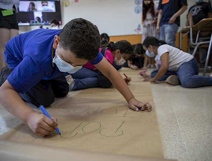 Primer plano de niño escribiendo sobre cartulina, en el suelo. En segundo plano hay otros niños y niñas.